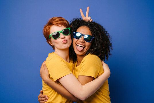Zwei diverse junge Frauen in gelben Shirts mit Sonnenbrillen umarmen sich vor blauer Wand &ndash; Freundschaft Lifestyle Diversit&auml;t