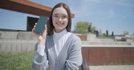 A hand-held portrait of a young female student in casual clothes wearing glasses, sitting on a concrete bench in an urban park and using a phone. © Anatoly Repin