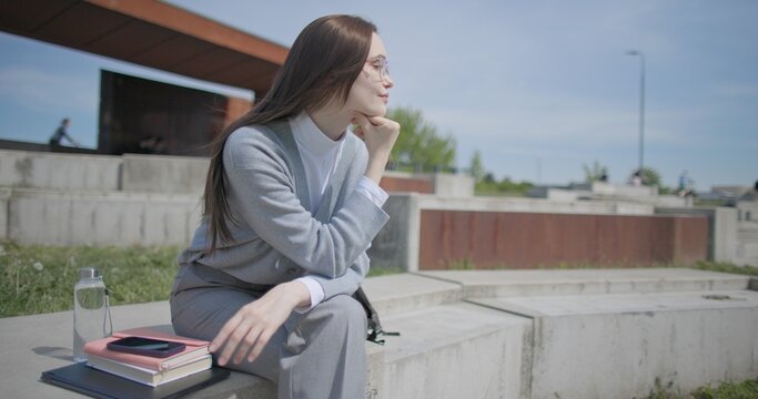 A handheld shot of a young female student in casual clothes sitting on a concrete bench in an urban park, enjoying a sunny summer day.