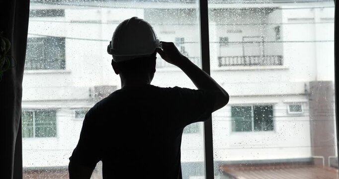 A silhouette of a male engineer stands inside a house, watching the rain fall through a window. The engineer is wearing a safety helmet.