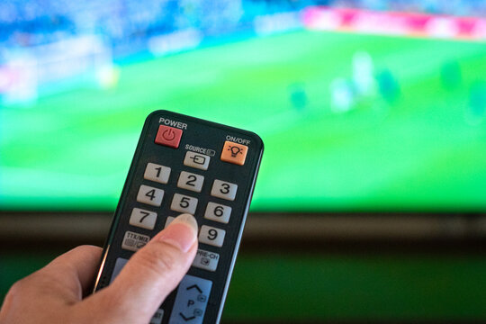 Hand Holding Remote Control Pointing at Television showing football match in action.