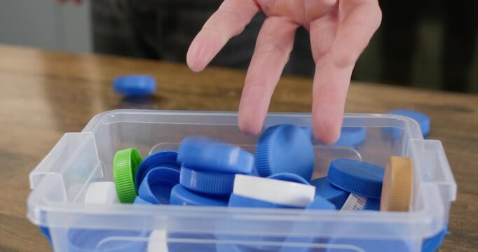 Adult hand releases blue bottle caps into clear sorting box on wooden table. Plastic lids fall into container supporting recycling process through careful separation