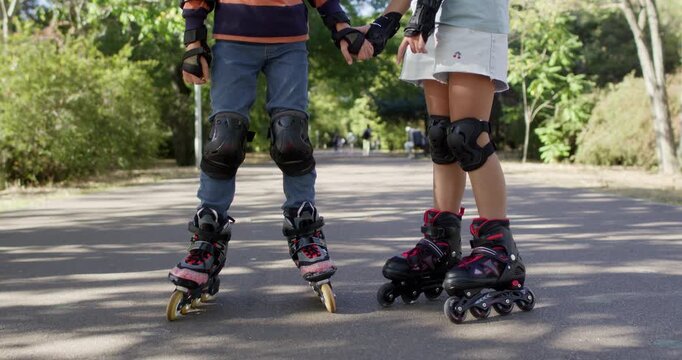 Girl supports brother on rollerblades while standing on park path. Children in helmets hold hands practicing balance together during slow outdoor skating lesson