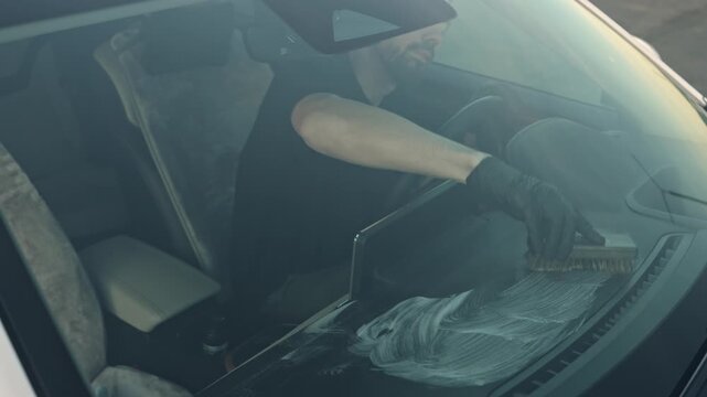 Man wipes the dashboard of a car with a cloth, looking outside the cabin through the windshield