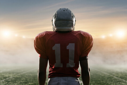 Player stands on field, back to camera. Helmet gleams, jersey reads "11". Dusk casts golden glow over scene. Stadium lights shine far in background. Mist drifts across grassy surface