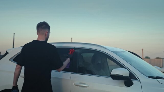 Man Spraying And Wiping Car Window Exterior, Open Lot Backdrop, Concentrated Sweep Across Glass