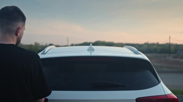 Man Sprays Rear Windshield With Cleaner At Dusk, Homeowner Wipes Hatchback Glass While Sunset Casts Warm