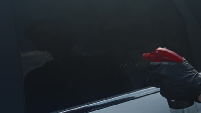 Auto Detailer Cleaning Tinted Car Window With Blue Microfiber Cloth And Red Spray Bottle, Gloved Hand Removes