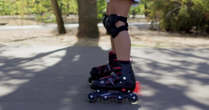 Girl skates along sunny green park pathway with helmet and elbow pads. Child leans forward slightly keeping rollerblade motion controlled during active park practice