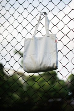 White tote bag hangs on chain-link fence. Soft lighting highlights fabric texture and simple design. Background shows grassy field and blurred trees. Casual