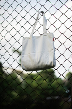 White tote bag hangs on chain-link fence. Soft lighting highlights fabric texture and simple design. Background shows grassy field and blurred trees. Casual