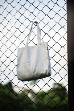 White tote bag hangs on chain-link fence. Soft sunlight highlights fabric's gentle folds. Background features blurred green grass and distant trees. No people visible, suggesting quiet, empty scene