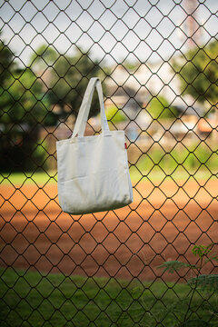 Simple cotton tote hangs on chain-link fence. Sunlight glows behind green trees and grass. Tote shows soft folds and clean lines. Natural scene suggests casual, everyday use