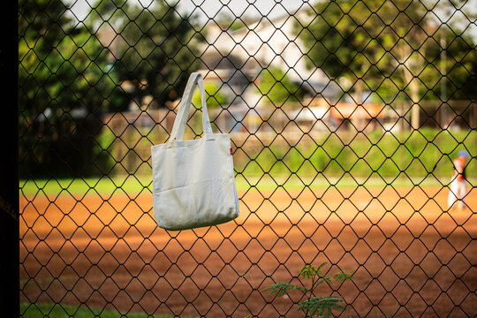 Simple cotton tote hangs on chain-link fence. Sunlight glows behind green trees and grass. Tote shows soft folds and clean lines. Natural scene suggests casual, everyday use