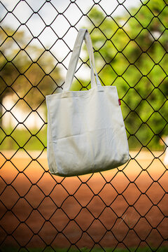 Simple cotton tote hangs on chain-link fence. Sunlight glows behind green trees and grass. Tote shows soft folds and clean lines. Natural scene suggests casual, everyday use