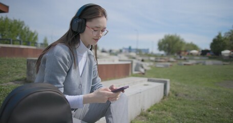 A hand-held shot of a young female student in casual clothes, wearing glasses and headphones, sitting on a concrete in an urban park on a sunny summer day and listening to music from a mobile phone © Anatoly Repin