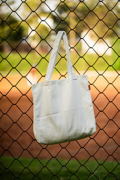 White tote bag hangs on chain-link fence. Blurry background shows green grass and dirt field. Child in distance wears cap and holds bat. Sunlit scene suggests casual park or baseball setting