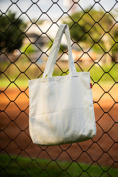 Simple tote bag hangs on chain-link fence. Soft fabric shows subtle wear and natural folds. Background holds green trees and blurred backyard. Scene suggests casual, everyday outdoor setting