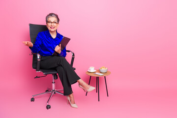 Fototapeta premium Confident senior businesswoman in blue blouse sits in pink studio with coffee and pastry promoting professional lifestyle