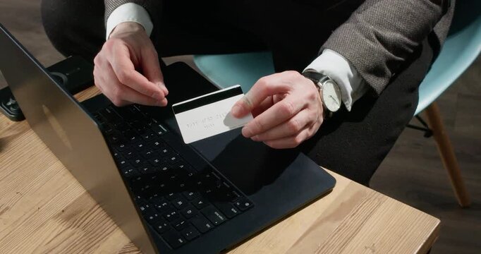Online scammer holds bank card above laptop during payment fraud setup. Man studies card details positioning hands over keyboard for deceptive transaction slow motion
