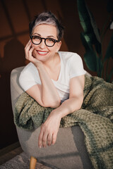 Fototapeta premium Smiling mature woman with glasses relaxing in an armchair wrapped in a green blanket, cozy home portrait conveying warmth, confidence and contentment