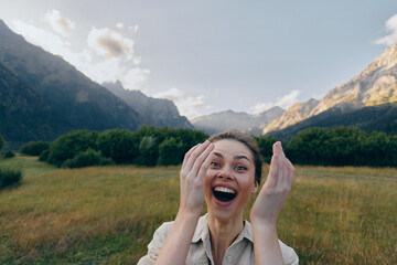 Fototapeta premium A young woman with brunette hair and a beige shirt expresses joy and excitement in a mountain meadow under a clear sky during daytime.