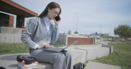 A hand-held shot of a young female student in casual clothes wearing glasses sitting on a concrete bench in an urban park on a sunny summer day, working on a laptop and typing on a keyboard. © Anatoly Repin