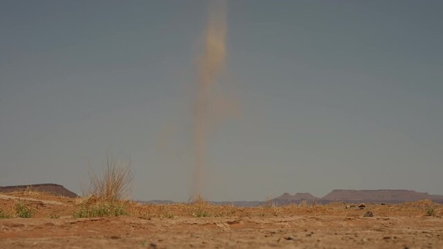 Tall dust devil whirlwind rising from dry desert floor against hazy sky with rocky mesas in Morocco