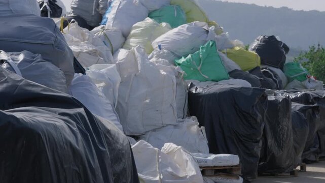 Rows of bulk plastic bags and covered pallets storing collected waste outside a recycling plant yard