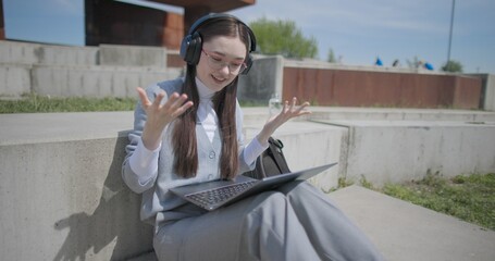 A hand-held shot of a young female student in casual clothes, glasses, and headphones making a video call on a laptop while sitting on a concrete bench in an urban park. © Anatoly Repin