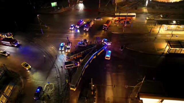 Aerial night view of a busy urban traffic intersection with emergency vehicles, buses, cars, wet reflective pavement, and a brightly lit circular plaza in the background
