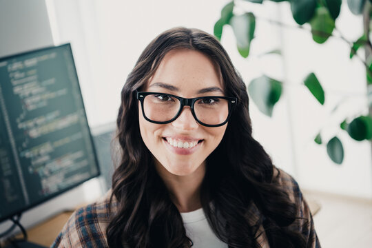 Smiling female software developer wearing glasses working on code in a bright home office with monitor and plant, casual modern portrait conveying remote work and productivity