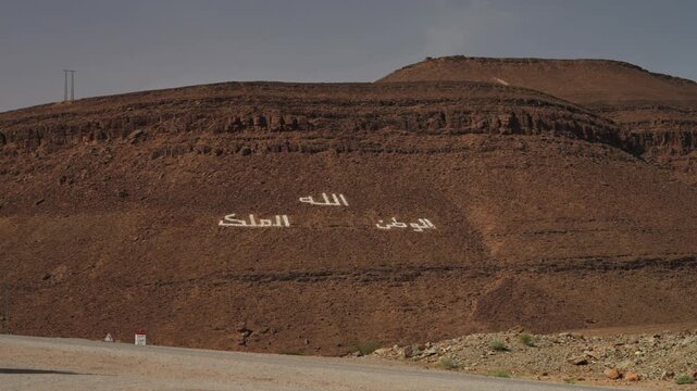 White Arabic motto God Homeland King painted on steep rocky hillside along desert road in Morocco