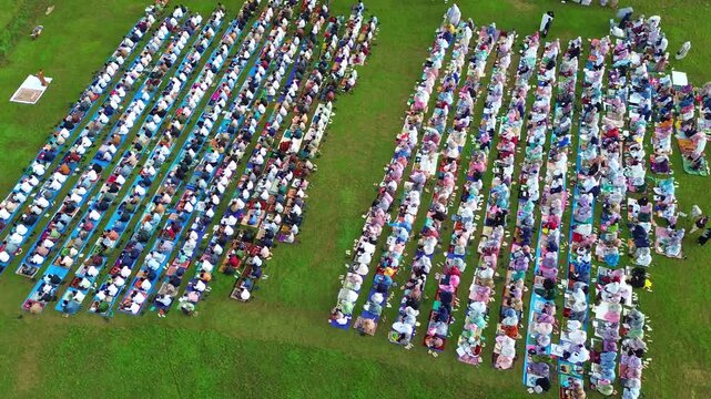 Drone shot of Muslim congregation praying Eid prayer outdoors, showcasing unity and faith on green field.
