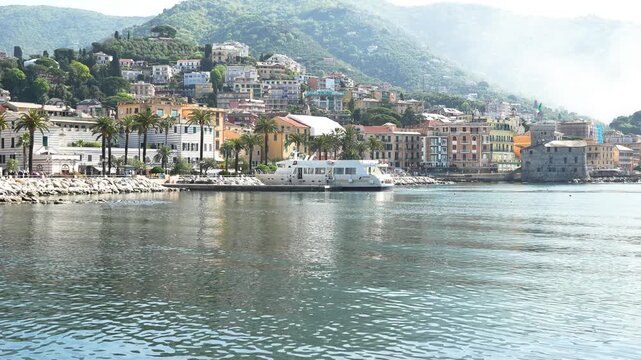 Wide angle static shot of Rapallo&rsquo;s picturesque seafront, featuring colorful Ligurian buildings, palm-lined promenade, calm harbor waters, and lush green hills under bright daylight.