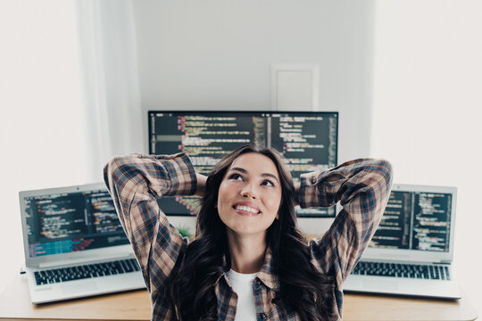 Relaxed female developer smiling at her home office with multiple laptops and code on screens, enjoying a moment of inspiration and productivity while working remotely on software development