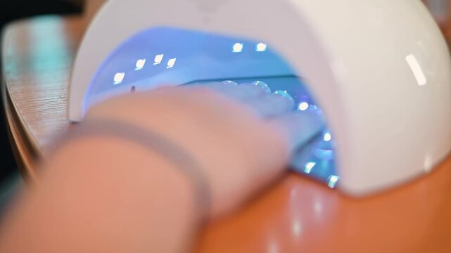 A hand is placed inside a UV nail dryer, showing the drying process of manicure treatments. Soft blue lights illuminate the area as the hand remains still, capturing the application of nail polish