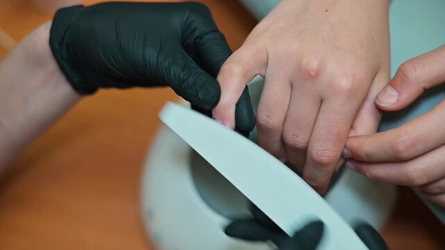 A nail technician performs a filing procedure on a client's nails in a beauty salon. The technician, wearing black gloves, focuses on shaping the nails while ensuring precision