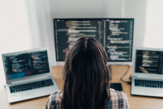 Female programmer working on multiple screens coding at a home desk with laptop and monitor for web development and software engineering