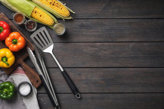 BBQ tools and fresh vegetables flat lay on dark wooden surface with copy space