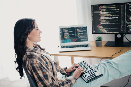 Young woman programmer coding from a relaxed home office with laptop and dual monitors, smiling while typing on a keyboard in a casual flannel &mdash; remote developer workspace