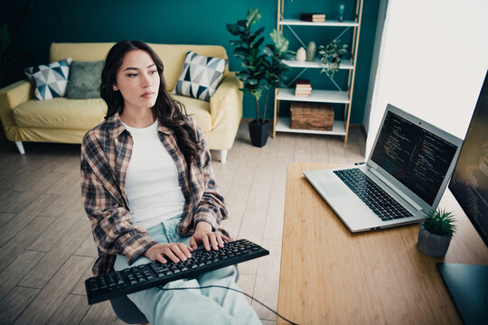 Young woman programming from home in a relaxed living room workspace, typing on an external keyboard with laptop displaying code, remote work in a casual modern home office