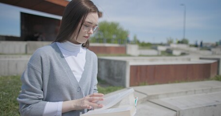 A hand-held shot of a young female student in casual clothes and glasses reading a book while flipping through pages while sitting in an urban park. © Anatoly Repin