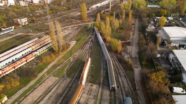High-angle drone perspective of a sprawling train storage yard with multiple parallel tracks, weathered passenger coaches, maintenance buildings, and nearby residential blocks under soft sunlight