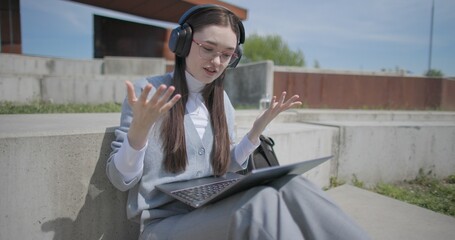 A hand-held shot of a young female student in casual clothes, glasses, and headphones making a video call on a laptop while sitting on a concrete bench in an urban park. © Anatoly Repin