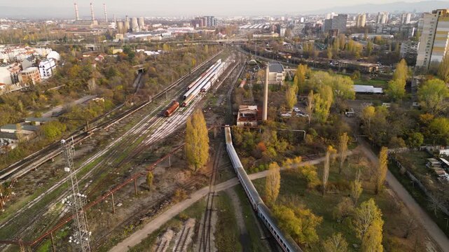 High-angle drone perspective of a sprawling train storage yard with multiple parallel tracks, weathered passenger coaches, maintenance buildings, and nearby residential blocks under soft sunlight