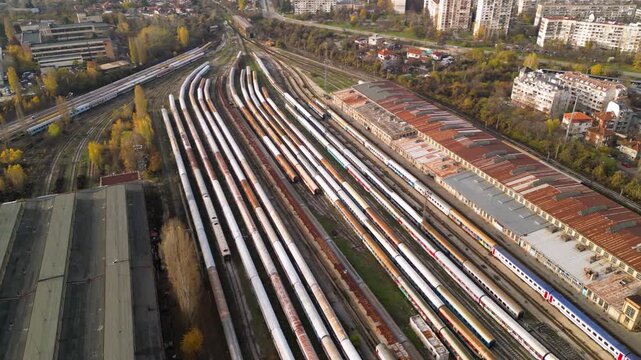 High-angle drone perspective of a sprawling train storage yard with multiple parallel tracks, weathered passenger coaches, maintenance buildings, and nearby residential blocks under soft sunlight