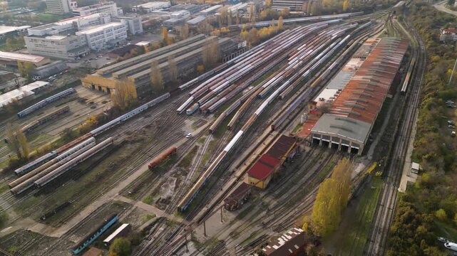 High-angle drone perspective of a sprawling train storage yard with multiple parallel tracks, weathered passenger coaches, maintenance buildings, and nearby residential blocks under soft sunlight