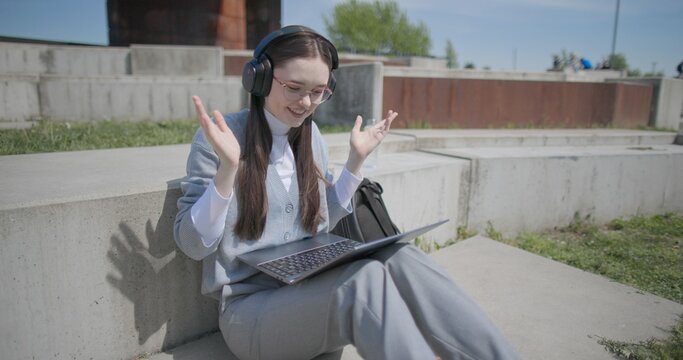 A hand-held shot of a young female student in casual clothes, glasses, and headphones making a video call on a laptop while sitting on a concrete bench in an urban park.