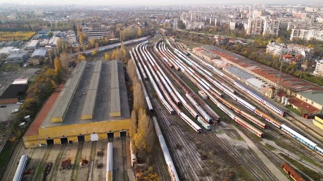 Aerial view of a large railway depot with long rows of parked passenger trains between industrial sheds and city neighborhoods, captured in warm late-afternoon light